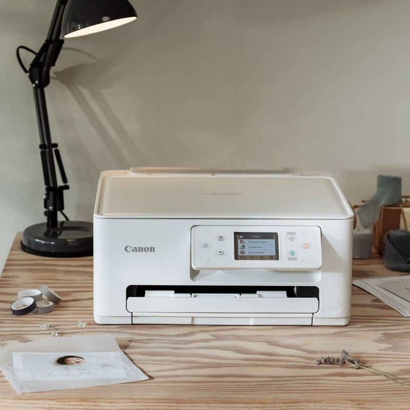 Canon Drucker auf einem Holztisch mit Fotodrucken, Bastelzubehör und einer Schreibtischlampe in einem kreativen Arbeitsbereich.