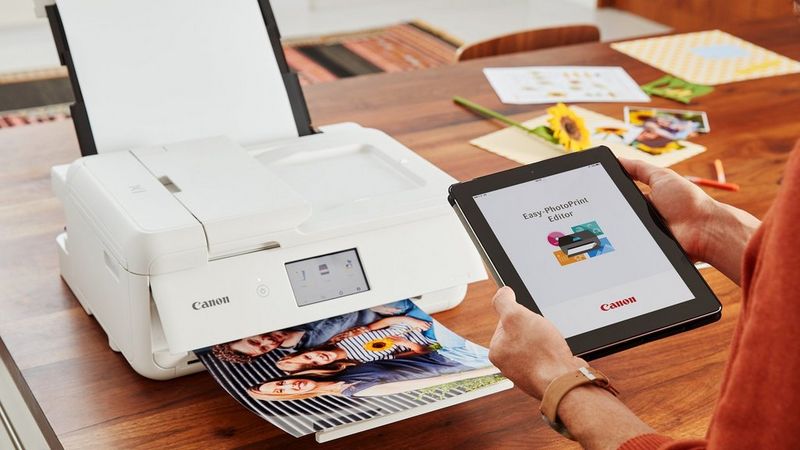 Auf einem Tablet ist der Easy-PhotoPrint Editor geöffnet; daneben druckt ein Canon Drucker auf einem Schreibtisch ein Foto aus.