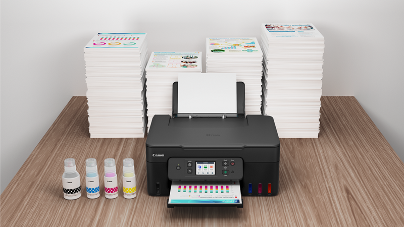 Auf einem Schreibtisch mit Tintenflaschen und Papierstapeln steht ein Drucker der PIXMA G Serie (MegaTank), aus dem ein Farbdruck kommt.