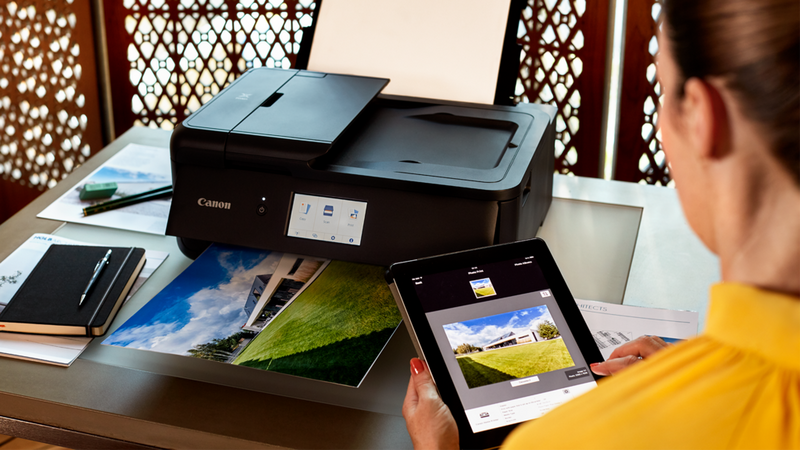 Auf einem Tablet ist die Canon PRINT App geöffnet; daneben druckt ein Canon Drucker auf einem Schreibtisch ein Foto aus.