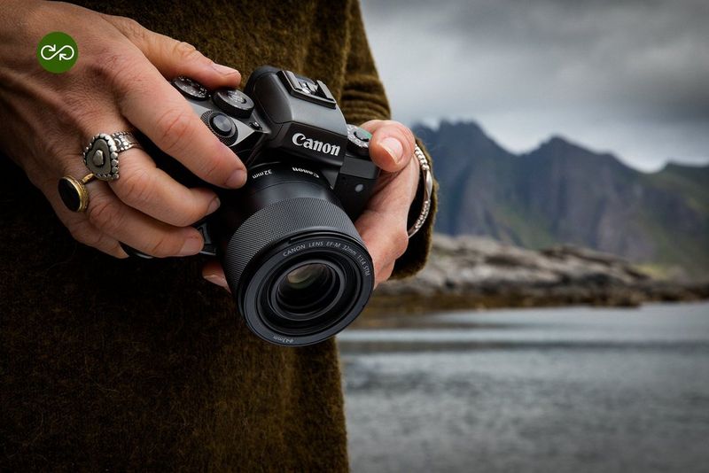 Eine Person hält eine spiegellose Canon Kamera in der Hand; daneben ein See, mit Bergen im Hintergrund