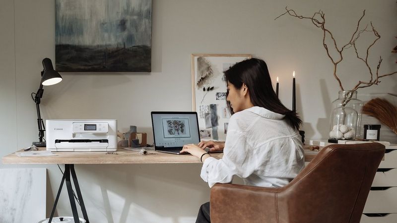 Person, die an einem Laptop neben einem Canon-Drucker im Homeoffice mit Grafiken und Deko arbeitet.