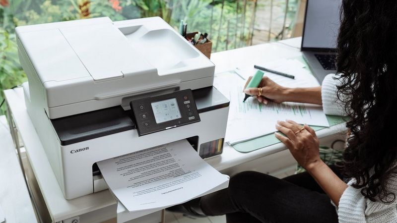 Canon MAXIFY GX2051 Drucker neben einer Person, die im Homeoffice in ein Notebook schreibt.