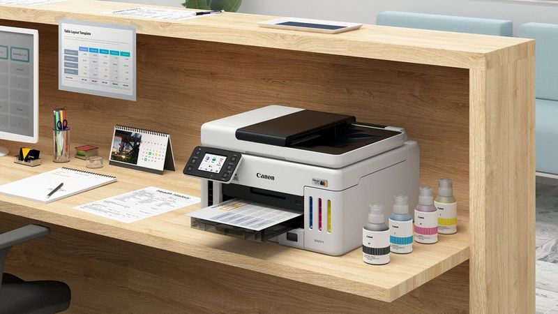 Canon MAXIFY GX6150 Drucker auf Holzarbeitsplatz beim Drucken von Dokumenten neben Tintenflaschen und Schreibwaren.