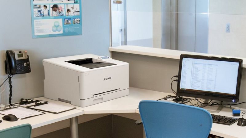Canon i-SENSYS LBP647Cdw Drucker auf einem Schreibtisch neben einem Computermonitor.