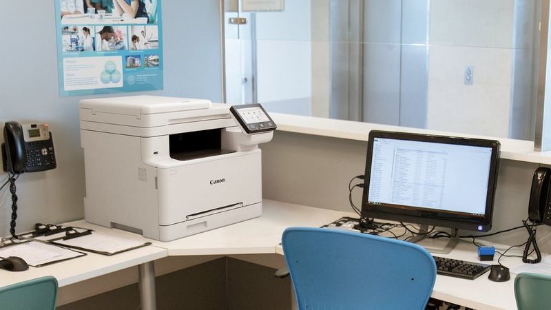 Canon i-SENSYS MF664Cdw Drucker auf einem Schreibtisch neben einem Computermonitor.