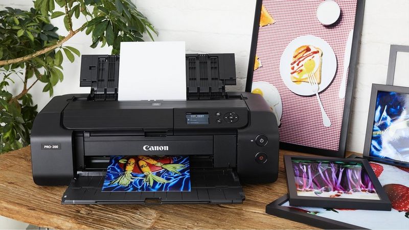 Canon PIXMA PRO-200S Drucker beim Drucken von farbenfrohen Grafiken neben gerahmten Ausdrucken und einem Tablet.
