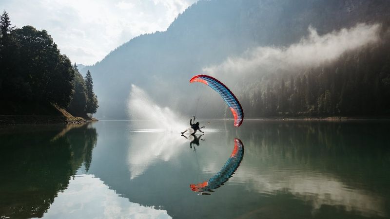 Ein Gleitschirmflieger über einem ruhigen Bergsee, darunter Sprühnebel und eine farbenfrohe Spiegelung