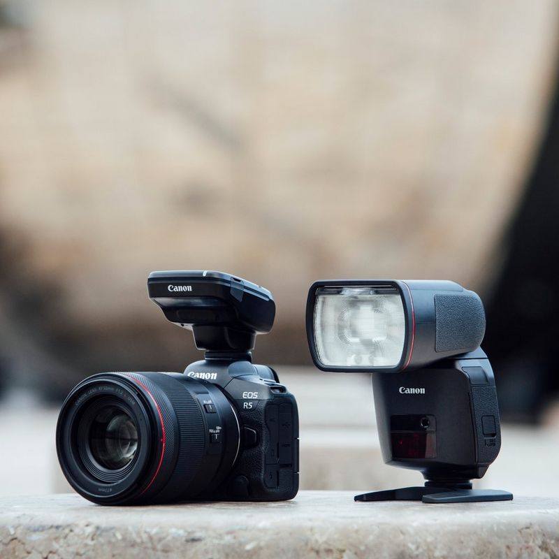 A Canon Speedlite EL-1 flash stands on a stone surface alongside a Canon EOS R5 camera with a Speedlite Transmitter ST-E3-RT in its Multi-function shoe.
