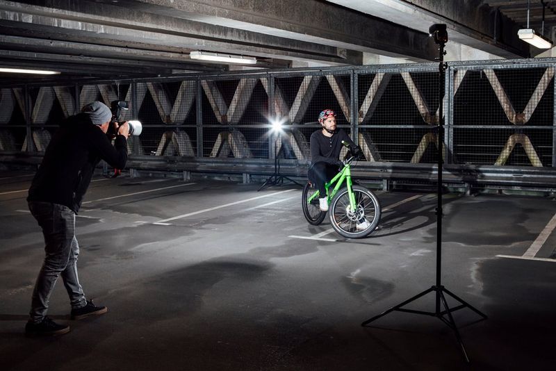 A man photographs a male model sitting on a mountain bike in a dark car park with a Canon Speedlite EL-1 flashgun.