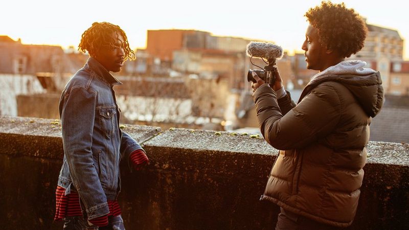 Two young men face each other, as they stand by a cement wall. One films the other using a small camera with a fluffy wind muffed microphone attached.