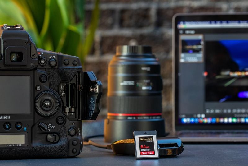 Een SanDisk Extreme PRO CFexpress-kaart leunt tegen een kaartlezer naast een Canon EOS-1D X Mark III, met een teleobjectief en een laptop op de achtergrond.