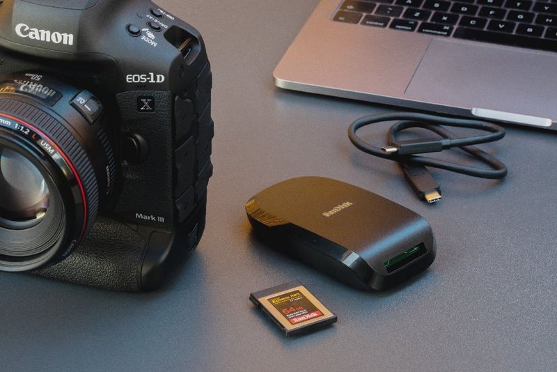 Een Canon EOS-1D X Mark III-camera ligt op een bureau naast een laptop en een SanDisk Extreme PRO CFexpress-geheugenkaart en -kaartlezer.