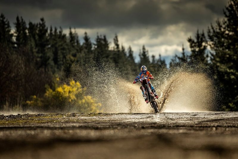 Een motorcrosser doet een wheelie door een modderpoel op een bospad. Het water spat op achter de motor.