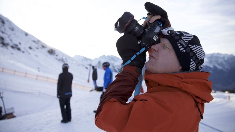 Fotograaf Richard Walch staat met een oranje jas en een zwarte gebreide muts in een besneeuwde bergomgeving en kijkt door de zoeker van de Canon EOS-1D X Mark II. Er zijn andere mensen op de achtergrond die winterkleding dragen en er zijn besneeuwde bergen zichtbaar onder een deels bewolkte lucht.