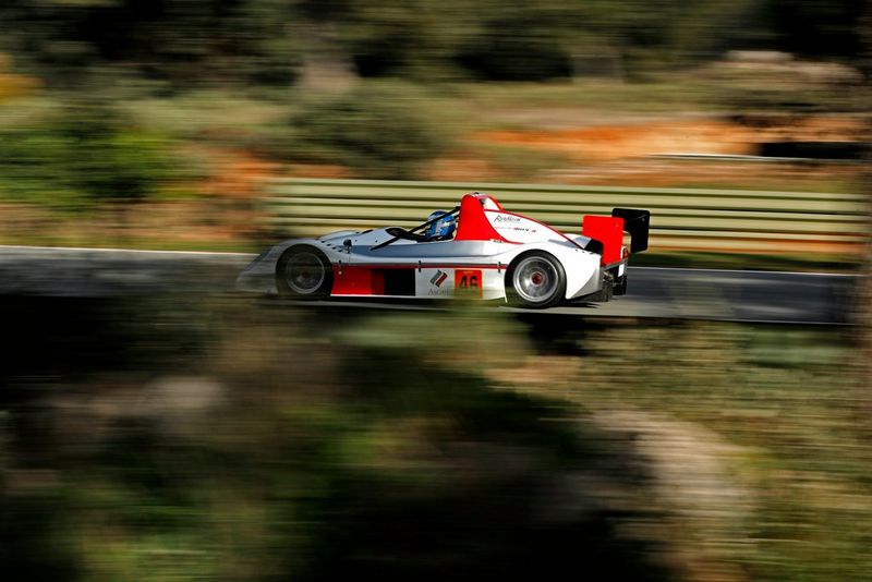 A fast-moving red and white racing car in sharp focus against a blurred background and foreground of greenery.