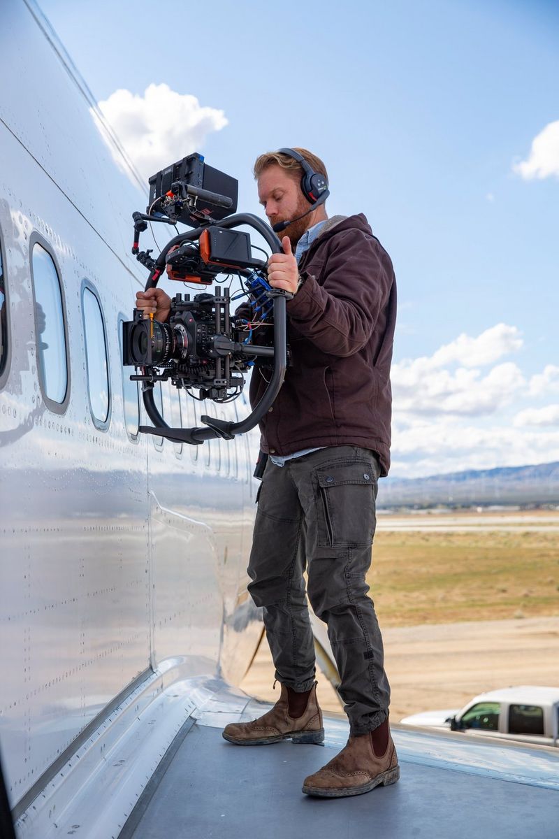 Cinematographer Steve Holleran holds a Canon EOS C300 Mark III in a gimbal as he stands on the wing of an aircraft filming inside through a window.