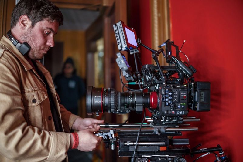 A film technician, wearing a tan jacket, adjusts a professional camera setup, the Canon EOS C400, in a room with red walls. He is focused on fine-tuning the equipment. 