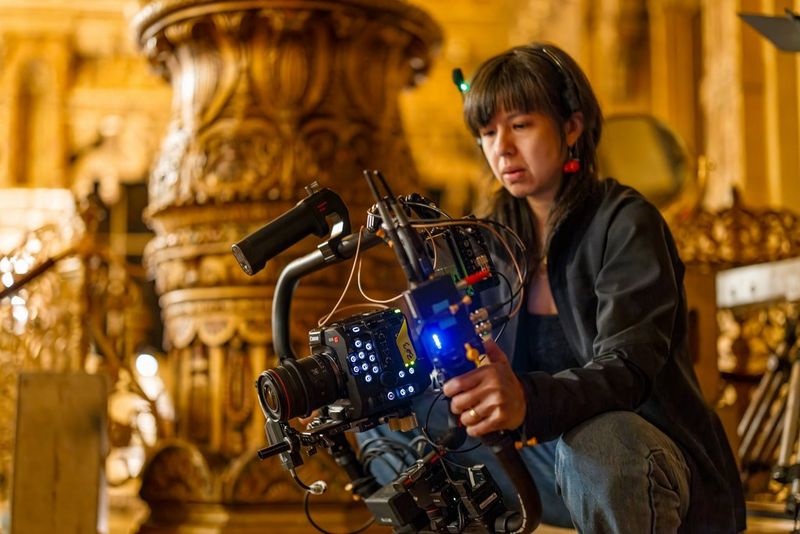  A close-up shot of a handheld Canon EOS C400 camera rig on a film set, with a focus on the detailed controls and wiring of the professional camera. The rig is positioned on a colourful carpet, and the hands of the cinematographer are visible.