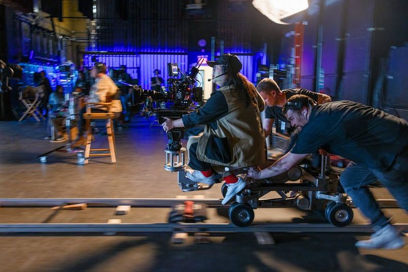 A camera operator sits on a dolly track, holding a professional camera setup, the Canon EOS C400, while a crew member pushes the dolly during a fast-paced film shoot. The scene is set in a bustling studio with various equipment and crew members visible in the background.