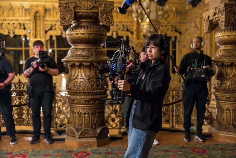 A cinematographer, dressed in dark clothing, operates a handheld Canon EOS C400 rig in a lavishly decorated venue. Other crew members, also in dark attire, are seen in the background, some holding additional camera equipment.