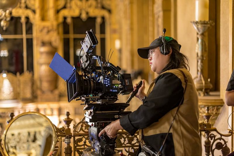 A filmmaker, wearing a black cap and tan jacket, operates a high-end Canon EOS C400 camera setup in an elaborate venue with ornate golden décor.