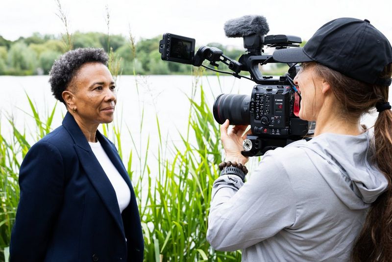 A filmmaker holds a Canon EOS C400 on her shoulder as she films a woman outdoors with a body of water in the background.