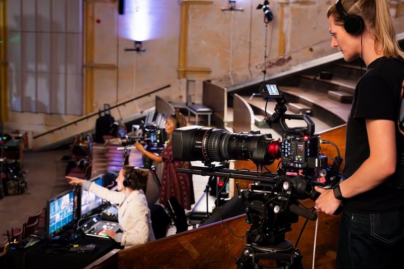 Een vrouw bedient een Canon EOS C400-camera op een statief in een grote aula, met andere camera's en de regisseur aan een control desk, met beeldschermen op de achtergrond. 