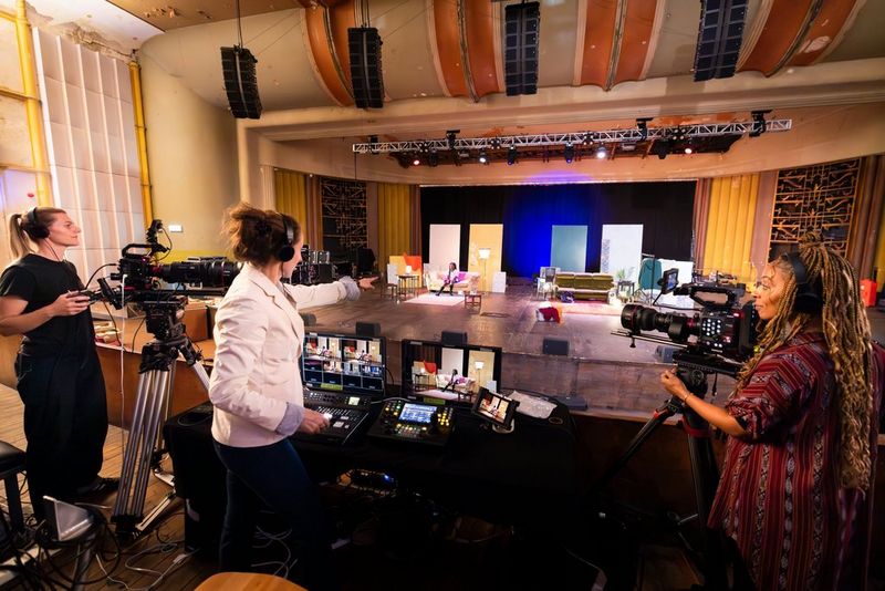 Sarah Thomas Moffat staat achter in een groot auditorium bij een control desk, en stuurt camera-operators aan die met Canon-filmcamera's het optreden op het podium filmen.