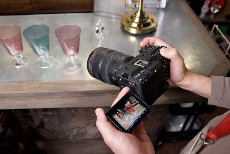 A user holds a Canon EOS C50 filming three coloured wine glasses on a table in an indoor setting.