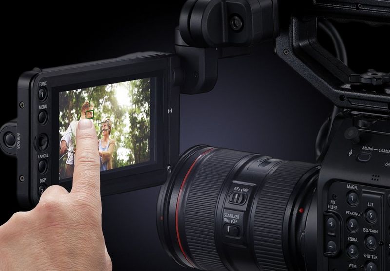 A user's finger taps a touchscreen attached to a Canon EOS C500 Mark II camera filming two people against a scene of trees backlit by bright sunshine.