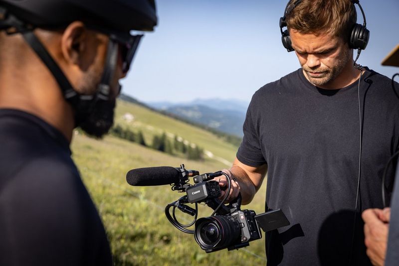 A filmmaker holding a Canon EOS C50, the screen folded out and a microphone attached to the camera's detachable handle.