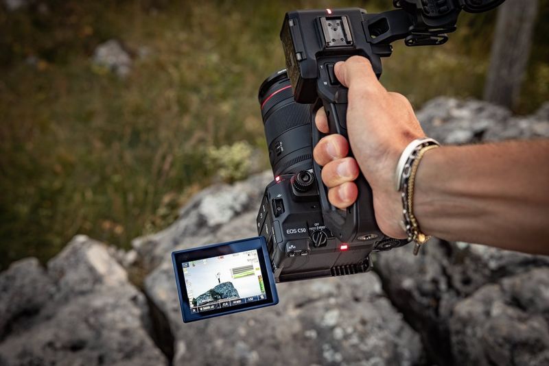 A user holds a Canon EOS C50 by the top handle, with the vari-angle screen showing a distant figure on a hilltop.