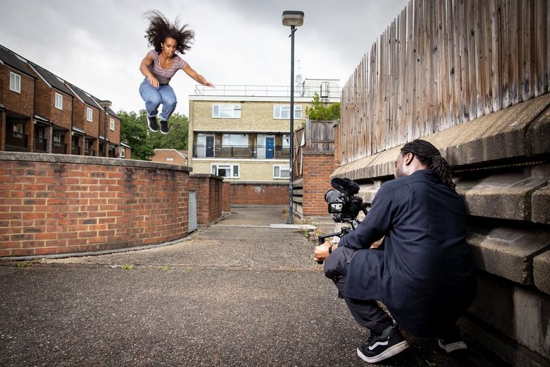 FIlmmaker Jolade Olusanya squats in an alleyway with a Canon EOS C70 in a gimbal to film a parkour athlete leaping over a brick wall.