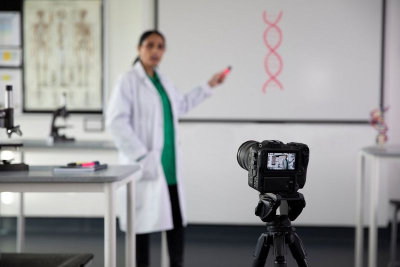 A presenter in a lab coat speaking to a Canon EOS C70 camera on a tripod.