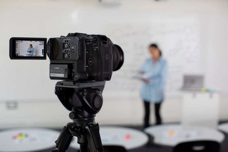 A Canon EOS C70 video camera on a tripod recording a person delivering a presentation in front of a whiteboard.