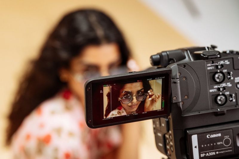 The LCD screen of a Canon EOS C80 camera displays a woman wearing sunglasses, looking directly at the lens while being filmed.