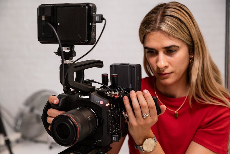 Cinematographer Carolina Nunes holding a Canon EOS C80 camera while looking at its LCD screen in a studio surrounding. 