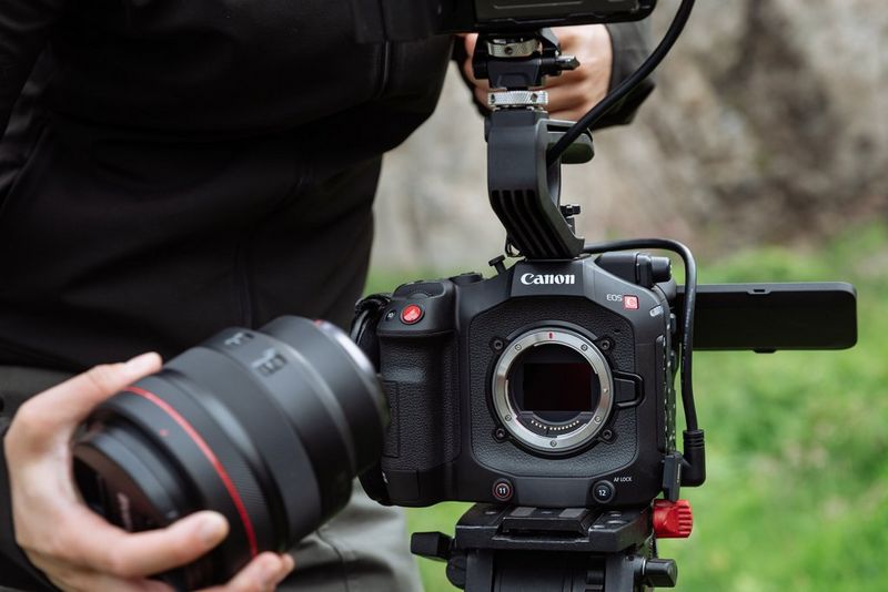 A hand poised to attach a Canon RF lens to a Canon EOS C80 camera mounted on a tripod.