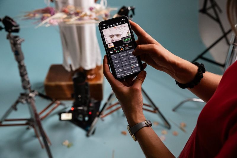 A hand holding a smartphone with the Canon Multi-Camera Control app  on screen with a blurred view of a studio setting with tripods in the background.