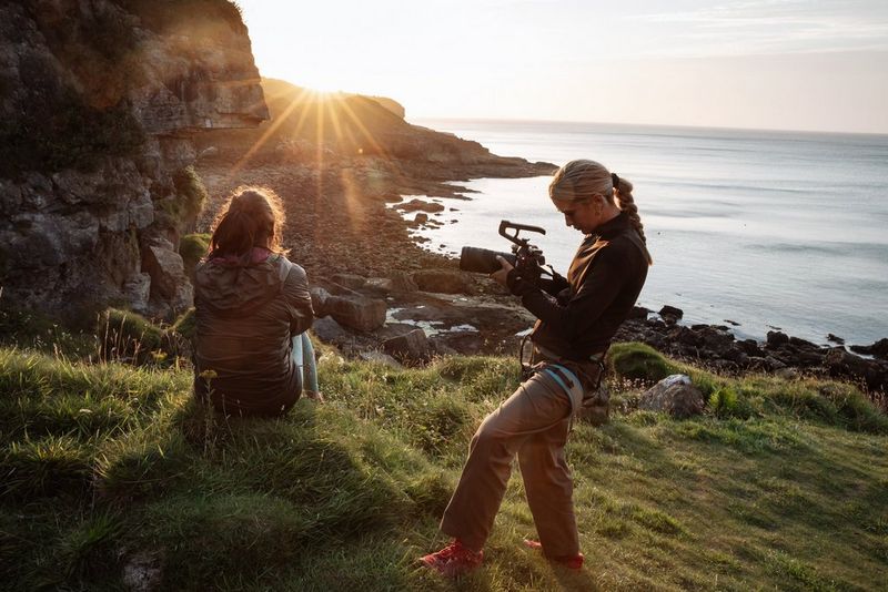 Cinematographer Carolina Nunes filming a person sitting on a grassy clifftop at sunset, overlooking the sea, using the Canon EOS C80 camera.