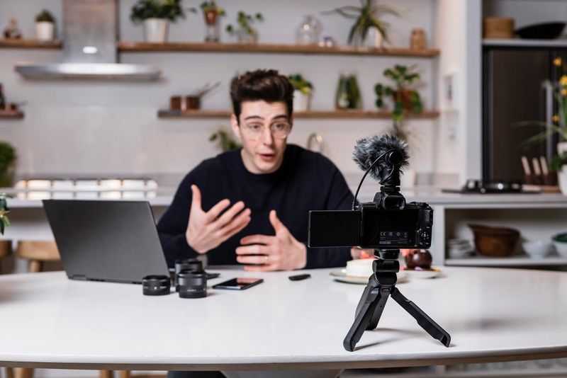 Мужчина сидит за столом с ноутбуком, телефоном и объективами для камер, пока его снимает камера Canon с установленным на ней микрофоном.