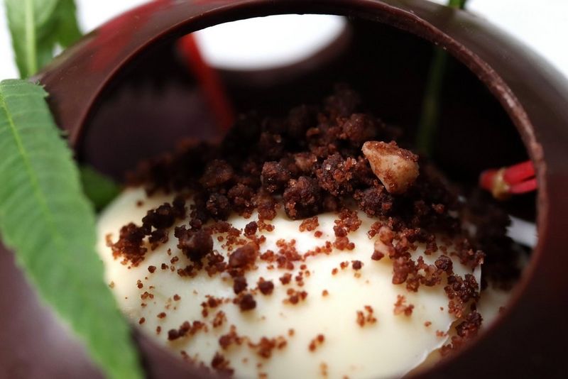 Крупный план шарика ванильного мороженого, покрытого тонким слоем шоколада и украшенного посыпкой, а также тонким зеленым листом.