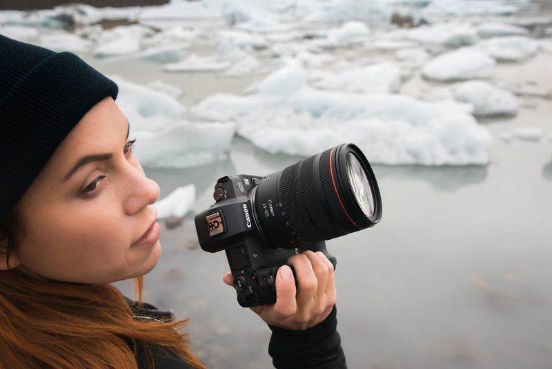 Una mano regge una fotocamera Canon a cui è collegato un obiettivo Canon RF 24-105mm F4 L IS USM. Sullo sfondo si vede un paesaggio ghiacciato con iceberg galleggianti su uno specchio d'acqua calmo.
