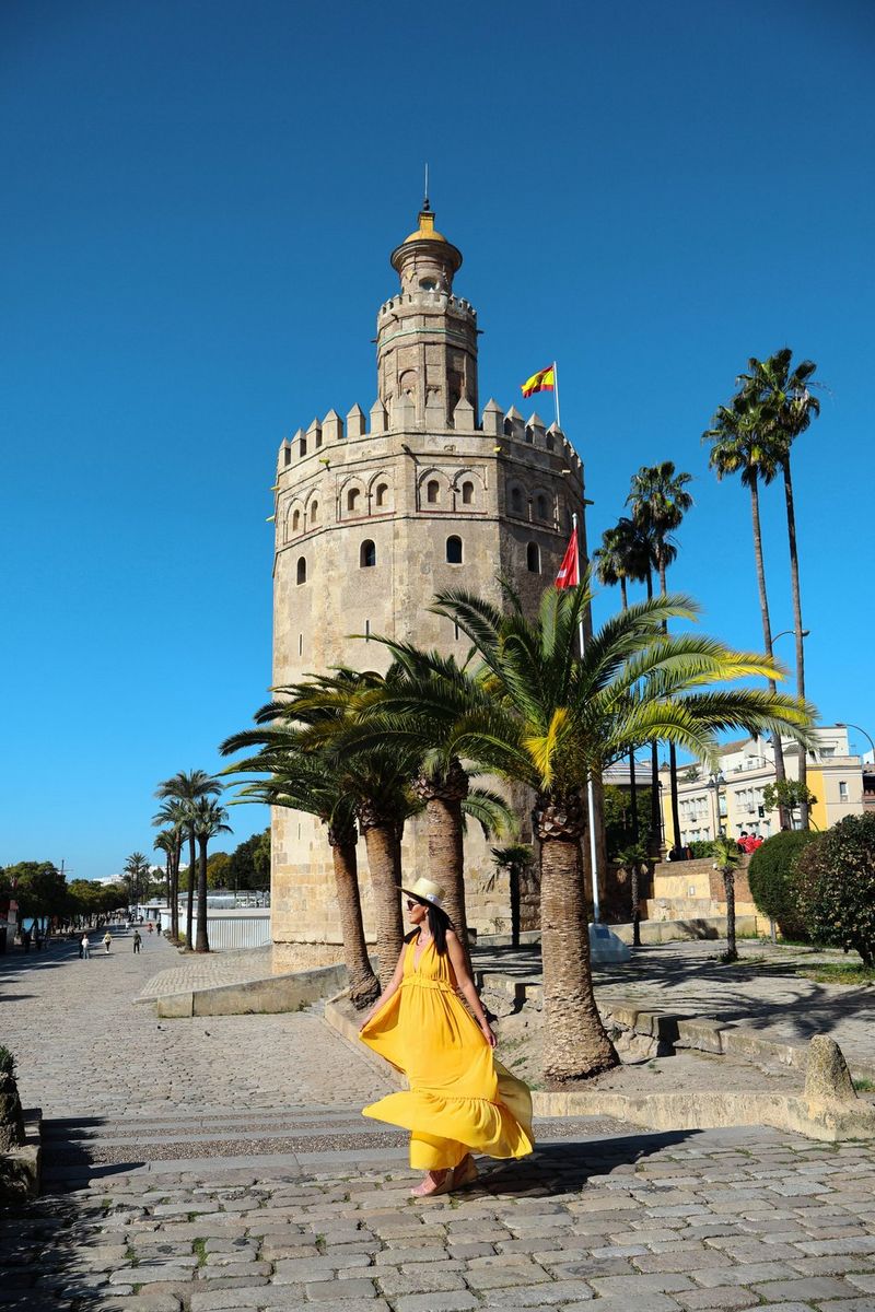 Une femme vêtue d'une robe jaune marche le long d'un sentier bordé de palmiers qui s'étend au loin, avec une tour en pierre en arrière-plan. Photo de voyage prise avec un objectif zoom Canon RF-S 18-45mm F4.5-6.3 IS STM.
