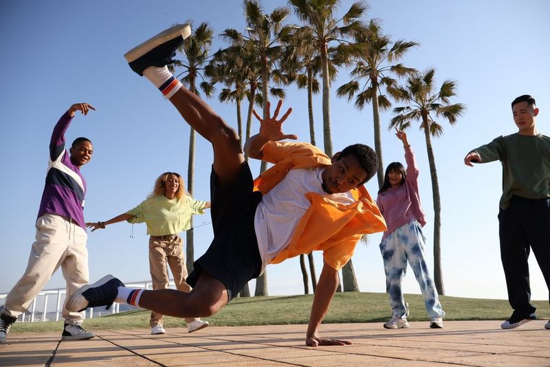 In a photo taken on a Canon EOS R10 with a high shutter speed, a street dancer performs a high kick with one hand on the ground. In the background are four other people and a clump of tall palm trees. 