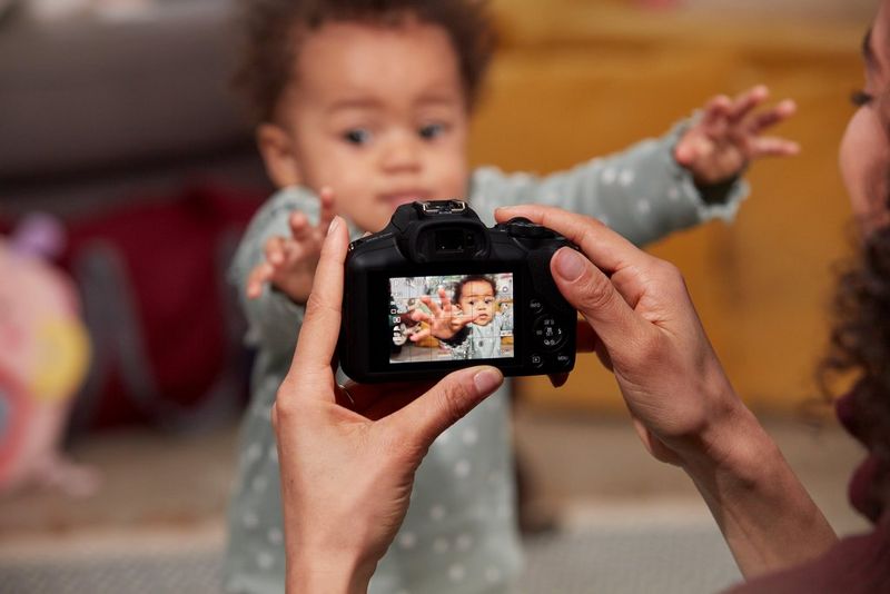 Человек держит в руках камеру Canon EOS R100 и фокусируется на ребенке, который идет в сторону камеры.