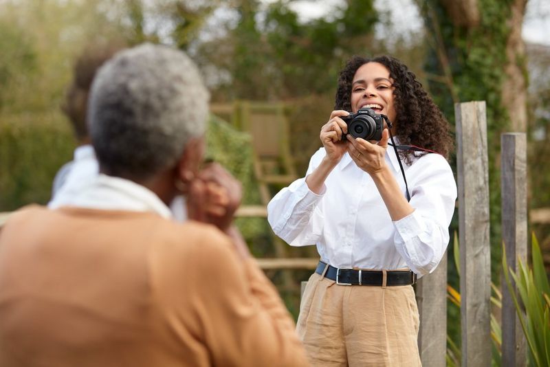 Une femme souriante portant une chemise blanche tient un Canon EOS R100 et le dirige vers deux personnes floues au premier plan, vues de dos.
