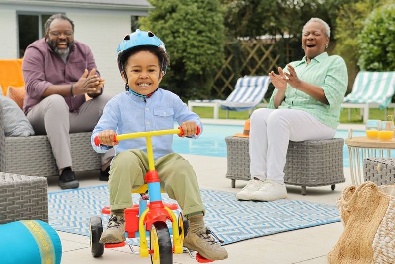 Улыбающийся ребенок проезжает на трехколесном велосипеде между смеющимися бабушкой и дедушкой, которые сидят возле открытого бассейна. Снято на камеру Canon EOS R100.
