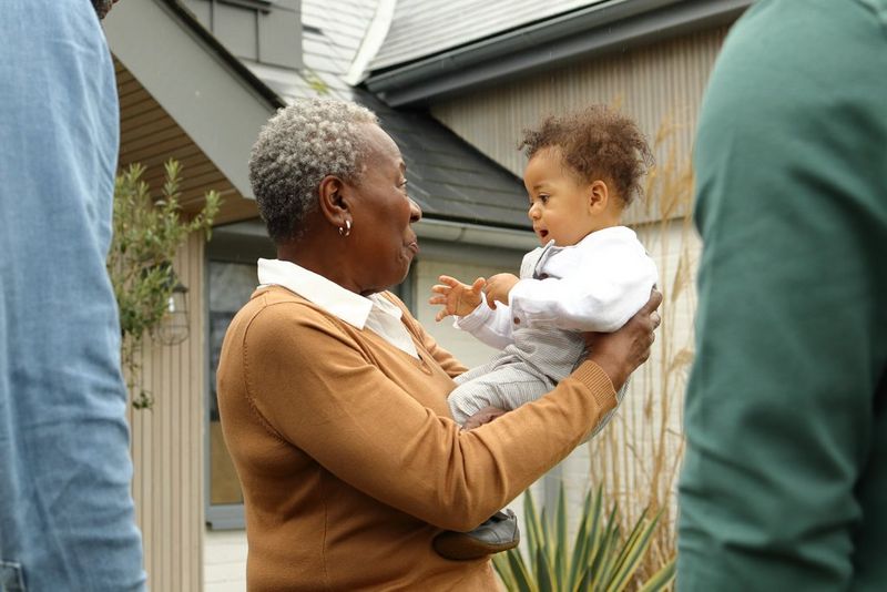 In a photo taken on a Canon EOS R100, a woman lifts a smiling child to her eye level in front of a house, framed by two people in the foreground.